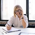 Smiling woman talking on the phone while writing notes at a desk in a modern office.